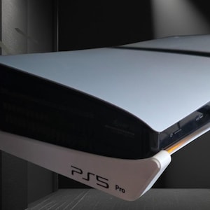 Puede incluir: Una consola PS5 Pro blanca y negra. La consola tiene un diseño curvo con una base blanca y una parte superior negra. El texto "PS5 Pro" está impreso en la base.