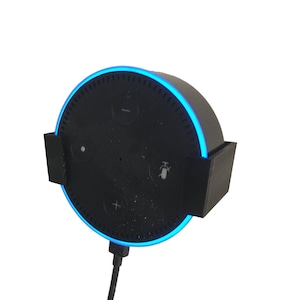 Pode incluir: Um alto-falante inteligente Amazon Echo Dot preto com um anel de luz azul ao redor da borda. O alto-falante está montado em uma parede com um suporte de plástico preto. O alto-falante está ligado com um cabo preto.