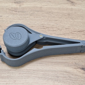 May include: A gray plastic coffee scoop with a built-in tamper. The scoop has a small handle and a larger, round base with a coffee bean design.