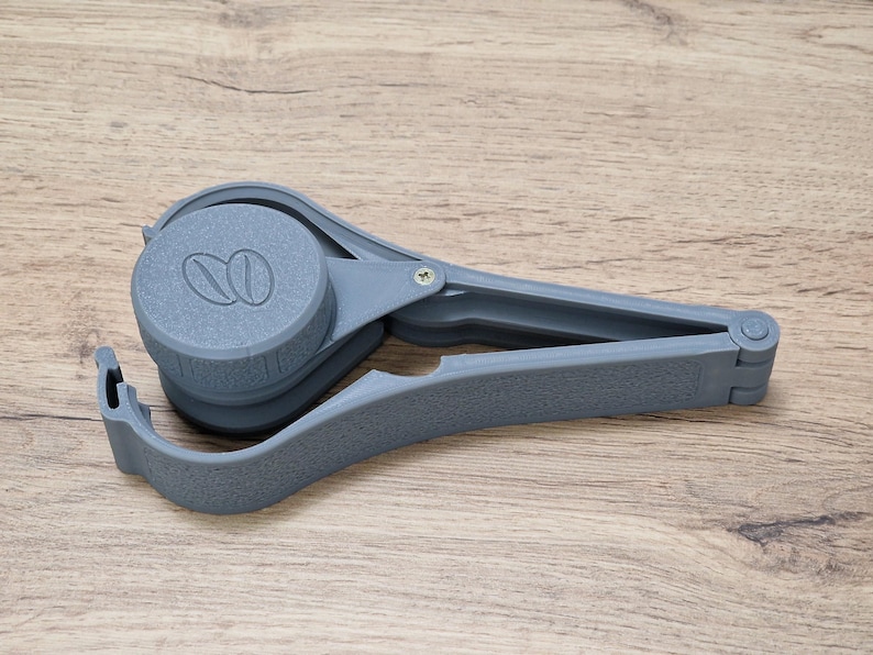 May include: A gray plastic coffee scoop with a built-in tamper. The scoop has a small handle and a larger, round base with a coffee bean design.