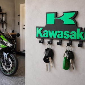 May include: A black and green Kawasaki key holder with five hooks, mounted on a grey wall. The Kawasaki logo and lettering are in green. Keys hang from the hooks. A green and black motorcycle is visible in the background.
