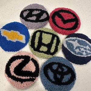May include: Seven round, colorful, crocheted coasters with different car logos. The logos include Hyundai, Mazda, Chevrolet, Honda, Ford, Lexus, and Volkswagen.
