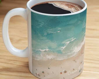 May include: A white ceramic mug featuring an aerial view of a sandy beach with turquoise ocean waves crashing on the shore. The design includes small figures on the sand and the text Ocean Breeze printed in a light script font near the bottom edge.