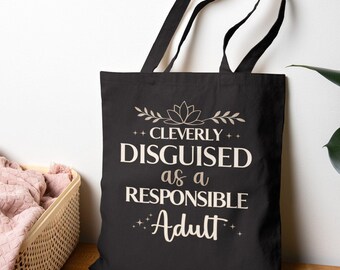 Może przedstawiać: Czarna torba typu tote z dwoma paskami na ramię, z białym nadrukiem "CLEVERLY DISGUISED as a RESPONSIBLE Adult" wyśrodkowanym na przodzie, uzupełnionym małym motywem kwiatowym nad tekstem oraz drobnymi akcentami w kształcie gwiazdek.