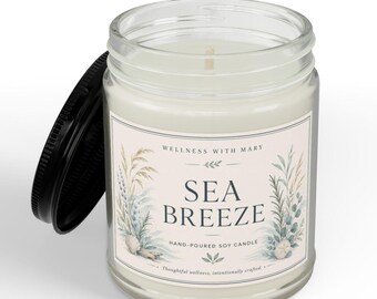 May include: A clear glass jar candle with a white wax fill and a black lid. The label features the text "WELLNESS WITH MARY", "SEA BREEZE", and "HAND-POURED SOY CANDLE" with illustrations of dried grasses, foliage, and small seashells on the sides.