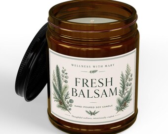May include: A brown glass jar candle with a black lid and a white label. The label features the text "WELLNESS WITH MARY", "FRESH BALSAM", and "HAND-POURED SOY CANDLE" alongside illustrations of green pine branches and small pinecones.