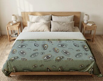May include: A sage green throw blanket featuring a repeating pattern of stylized eyes with dark outlines, gold accents, and long lashes. The blanket is draped over a bed with white bedding, two beige pillows, and two white pillows in a bedroom setting.