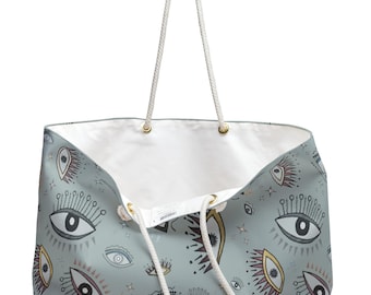 May include: A large tote bag with a light blue-gray exterior featuring a repeating pattern of stylized eyes with long lashes in black, white, and gold tones. The bag has a white interior lining and two long, white braided rope handles with gold grommets.
