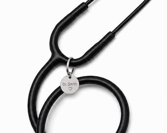 May include: A black medical stethoscope featuring a circular silver-toned metal charm attached to the tubing. The charm is engraved with the text Dr. Smith and a small heart symbol. The stethoscope has black ear tips and a black chest piece.