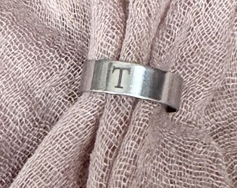 May include: A silver-toned metal ring featuring a flat band with the letter T engraved on the front. The ring is positioned against a textured, dusty pink fabric. A circular logo at the top reads MI Mary, and text at the bottom reads Professionally Engraved in My Studio.