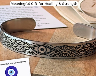 May include: A silver-toned metal cuff bangle featuring a continuous engraved pattern of multiple eyes. The bangle is accompanied by white informational cards with text including "Carry Intention. Attract Positivity." and a blue evil eye graphic.