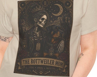 May include: A beige t-shirt featuring a dark, tarot-style graphic of a skeleton wearing a crown and leather jacket with a Rottweiler. The text reads THE ROTTWEILER MOM, LOYAL, STRONG, PROTECTIVE, with a crescent moon and stars in the background.