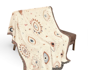 May include: A cream-colored woven throw blanket featuring a repeating pattern of stylized eyes in shades of terracotta, blue, and dark brown. The blanket has a textured knit construction and finished edges with short, multi-colored fringe detailing.