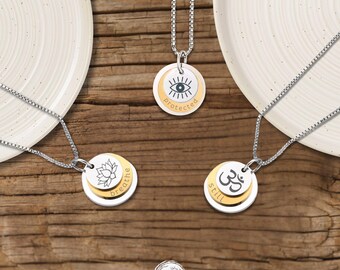May include: Three silver and gold-toned layered pendant necklaces on box chains. One pendant features a lotus flower with the word "breathe," another shows an eye symbol with "protected," and the third displays an Om symbol with "still."