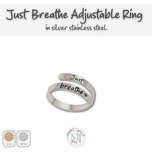 May include: A silver stainless steel adjustable ring with the words "Just breathe" engraved on it. The ring has a minimalist design and a heart symbol next to the word "breathe". The ring is shown on a white background.