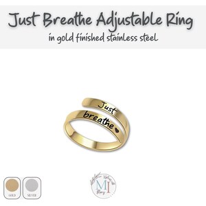 May include: Gold-finished stainless steel adjustable ring. The ring is engraved with the words "Just breathe" in black script. The ring has a heart symbol after the word "breathe". The text "Just Breathe Adjustable Ring" is at the top.