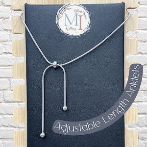 May include: A silver-toned snake chain anklet featuring an adjustable sliding bead mechanism and two dangling ends with small spherical terminals. The piece is displayed on a black card with the text Wellness Designs MI by Mary and Adjustable Length Anklets.