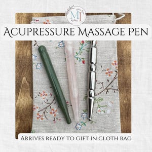 May include: An assortment of acupressure massage pens in green, pink, and silver, displayed on a floral patterned cloth bag. The text "ACUPRESSURE MASSAGE PEN" is above the pens. The text "ARRIVES READY TO GIFT IN CLOTH BAG" is below the pens.