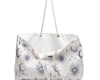 May include: A white tote bag featuring a repeating pattern of celestial motifs including suns, crescent moons, stars, and stylized eyes in shades of blue and grey. The bag has two long, white rope handles attached with gold-toned metal grommets.