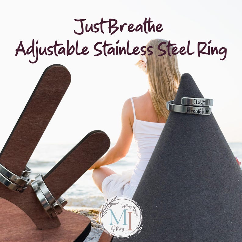May include: Stainless steel adjustable ring with the words "Just Breathe" engraved on it. The ring is displayed on a black cone-shaped stand. The background features a woman sitting near the ocean.