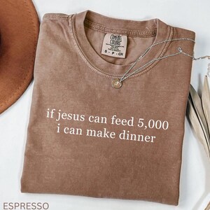 K&ouml;nnte beinhalten: Espressofarbenes T-Shirt mit dem wei&szlig;en Text "if jesus can feed 5,000 i can make dinner". Das Shirt hat einen Rundhalsausschnitt und ein Comfort Colors-Etikett. Eine silberne Halskette mit Anh&auml;nger liegt auf dem Shirt.