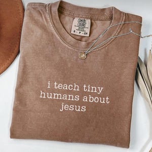 K&ouml;nnte beinhalten: Ein braunes Comfort Colors T-Shirt mit dem wei&szlig;en Text "i teach tiny humans about jesus". Eine silberne Halskette mit einem kleinen Anh&auml;nger liegt &uuml;ber dem Shirt. Ein brauner Hut ist in der oberen linken Ecke zu sehen.