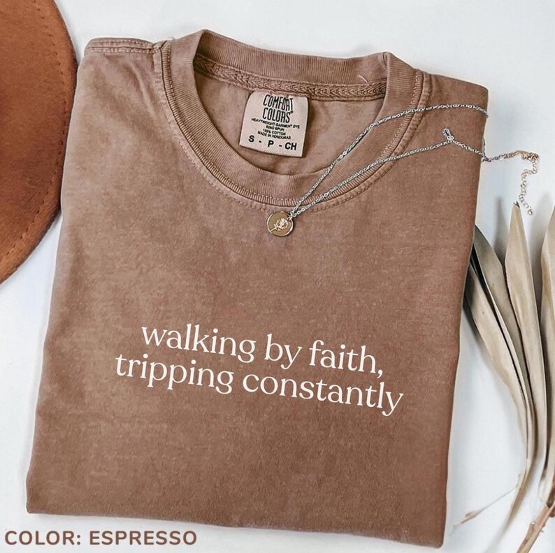 K&ouml;nnte beinhalten: Espressofarbenes Comfort Colors T-Shirt mit dem wei&szlig;en Text "walking by faith, tripping constantly". Eine silberne Halskette mit Anh&auml;nger liegt auf dem gefalteten Shirt.