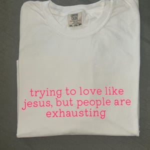 K&ouml;nnte beinhalten: Wei&szlig;es T-Shirt mit rosa Aufdruck "trying to love like jesus, but people are exhausting". Das gefaltete Shirt zeigt das Comfort Colors-Label. Ein l&auml;ssiges, bequemes Kleidungsst&uuml;ck.