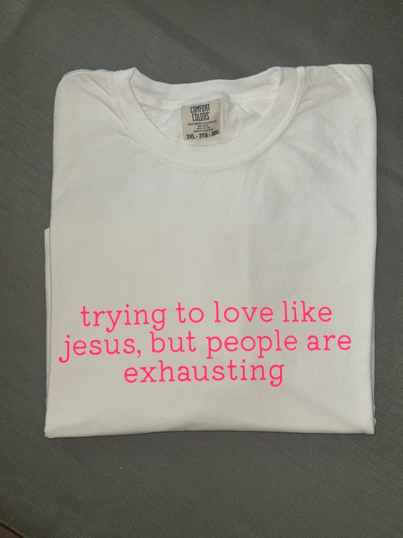K&ouml;nnte beinhalten: Wei&szlig;es T-Shirt mit rosa Aufdruck "trying to love like jesus, but people are exhausting". Das gefaltete Shirt zeigt das Comfort Colors-Label. Ein l&auml;ssiges, bequemes Kleidungsst&uuml;ck.