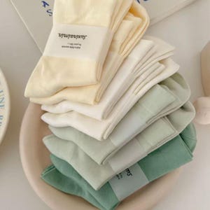 May include: A stack of folded socks in a gradient of colours ranging from pale yellow to cream, light sage green, and dark sage green. Each pair is secured with a white paper band featuring the text suxinaimin and Add a little warmth to your life.