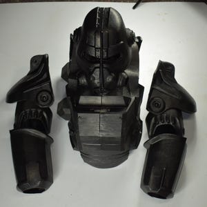 May include: A matte black power armor set consisting of a central chest piece with an integrated helmet and two separate arm components. The armor features a rigid, segmented design with mechanical detailing, including circular joints and vents.