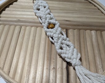 May include: Cream-colored macrame keychain with a wooden bead accent and a tassel. The keychain is attached to a bronze-colored clasp. The macrame is displayed on a light brown, round, wooden tray.