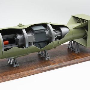 Könnte beinhalten: Detailliertes Modell einer Militärbombe in Olivgrün, mit einem Querschnitt, der die inneren Komponenten zeigt. Das Modell ist auf einem Holzsockel montiert und wird von grauen Ständern getragen. Die Bombe hat eine abgerundete Front und ein quadratisches Heck.