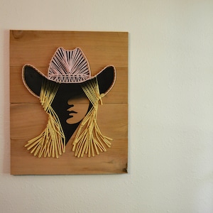 Puede incluir: Un adorno de pared de madera con un diseño de arte de hilo que representa a una mujer con un sombrero de vaquero rosa. El rostro de la mujer es una silueta negra y su cabello está hecho de hilo amarillo.