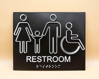 Può includere: Un cartello per i bagni in bianco e nero con un simbolo di famiglia e un simbolo di sedia a rotelle. Il cartello recita "RESTROOM" con il braille sotto.