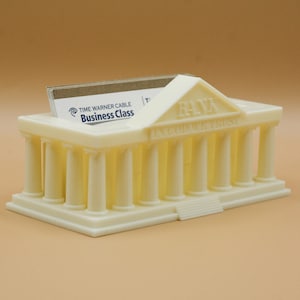 Op de afbeelding: Een witte plastic visitekaartjeshouder in de vorm van een miniatuurgebouw met kolommen en een fronton. Het gebouw heeft het woord "BANK" op de voorkant en de tekst "IN GOD WE TRUST" eronder. Een visitekaartje is zichtbaar in de houder, waarop "TIME WARNER CABLE Business Class" staat.