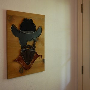 May include: A wooden wall art piece featuring a string art silhouette of a person wearing a cowboy hat. The silhouette is made with brown and black string and is surrounded by a brown wooden frame.
