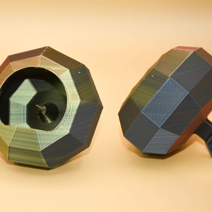 May include: Two geometrically shaped, 3D printed, black, green, and red spinning tops. One top is lying on its side, while the other is standing upright. The top standing upright has a small, black, circular base.