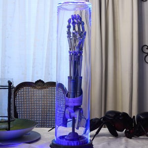 Puede incluir: Una mano robótica encerrada en un cilindro de vidrio transparente con una luz azul que ilumina la mano y una base con una fila de luces rojas.