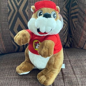 May include: A brown plush squirrel wearing a red shirt with a logo of a squirrel on it. The squirrel is wearing a red hat and has a big smile.