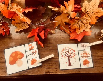 May include: Two illustrated cards with autumn themes, featuring a fox, mushrooms, and a tree with falling leaves. Wooden clothespins hold the cards. Artificial fall leaves in orange and red are scattered around the cards on a wooden surface.