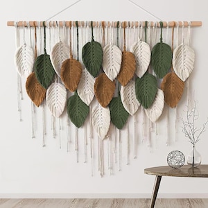 May include: A macrame wall hanging featuring a wooden dowel with hanging leaves in white, green, and brown. The leaves are arranged in rows, with long, dangling strings below. A decorative piece for home decor.