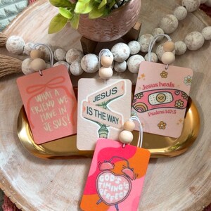 May include: A collection of pink and peach-colored rectangular ornaments with inspirational quotes and religious themes. Each ornament has a wooden bead accent and a string for hanging. The quotes include "What a friend we have in Jesus!" and "Jesus is the way."