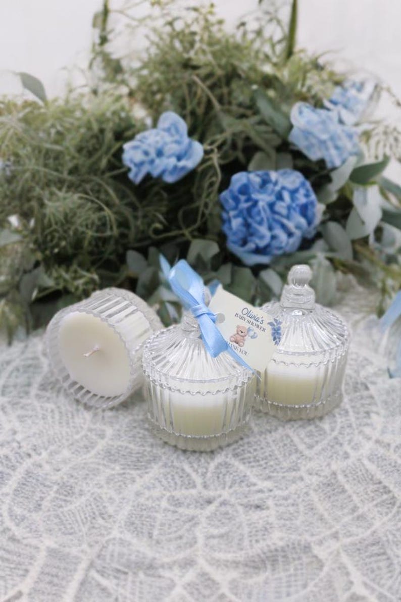 May include: Three small glass candle jars with white candles and blue ribbon accents. The jars have a domed lid and are sitting on a white crocheted surface. A tag with the text "Olivia's Baby Shower" is attached to one of the jars.