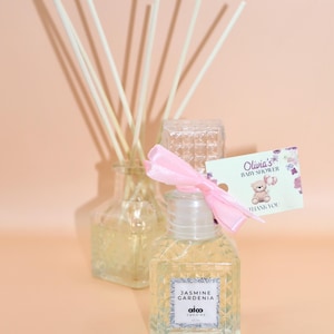 May include: A clear glass bottle with a pink ribbon and a thank you tag that says "Olivia's Baby Shower" with a cartoon bear. The bottle is filled with a clear liquid and has a label that says "Jasmine Gardenia" and "aico candles".