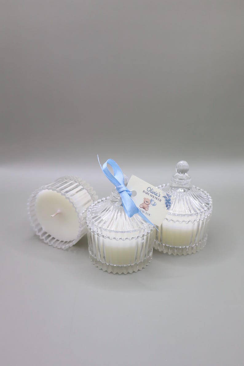 May include: Three clear glass candle jars with white candles. The center jar has a blue ribbon and a tag that reads "Olivia's Baby Shower Thank You".