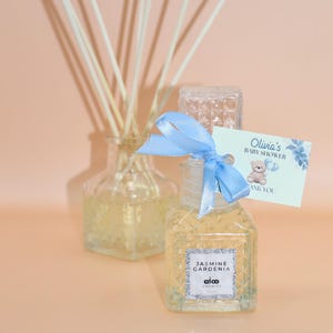 Puede incluir: Una botella de vidrio transparente con una cinta azul y una etiqueta que dice "Olivia's Baby Shower Thank You". La botella está llena de un líquido transparente y tiene una etiqueta que dice "Jasmine Gardenia alco candles".