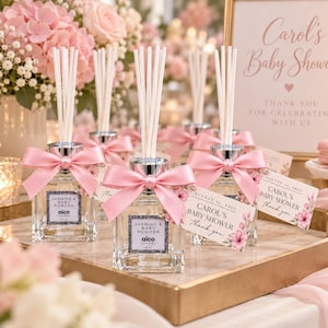 Könnte beinhalten: Quadratische Diffusorflaschen aus klarem Glas mit weißen Stäbchen, jeweils mit einer rosa Satinschleife verziert. Die Etiketten zeigen Jasmine & Baby Powder und Aico Candles 45 ml. Die angehängten Kärtchen tragen die Aufschrift Carol's Baby Shower.