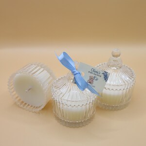 May include: Three clear glass candle jars with white candles inside. The jars have a fluted design and a lid with a knob on top. A blue ribbon is tied around one of the jars with a tag that says "Olivia's Baby Shower Thank You".