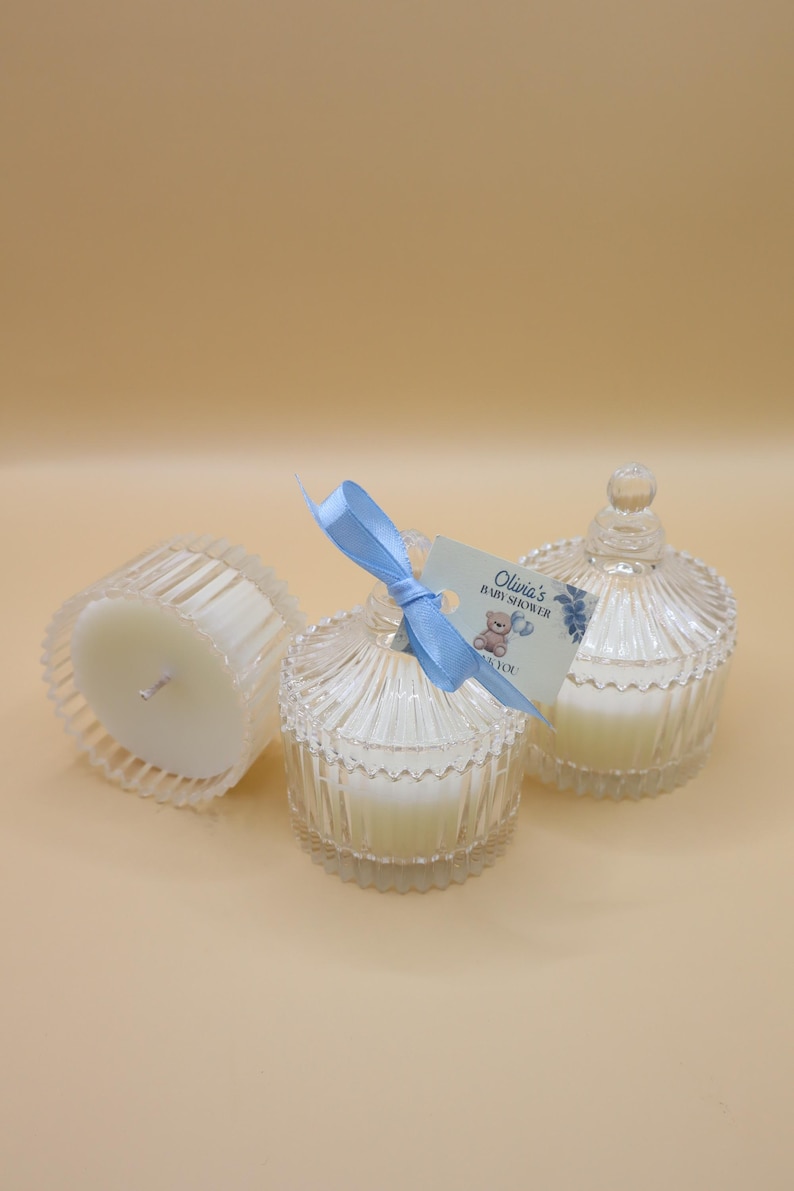 May include: Three clear glass candle jars with white candles inside. The jars have a fluted design and a lid with a knob on top. A blue ribbon is tied around one of the jars with a tag that says "Olivia's Baby Shower Thank You".